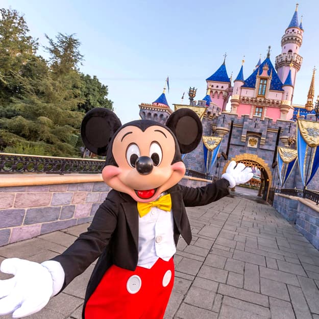 Mickey Mouse in tuxedo greets visitors in front of Sleeping Beauty Castle.
