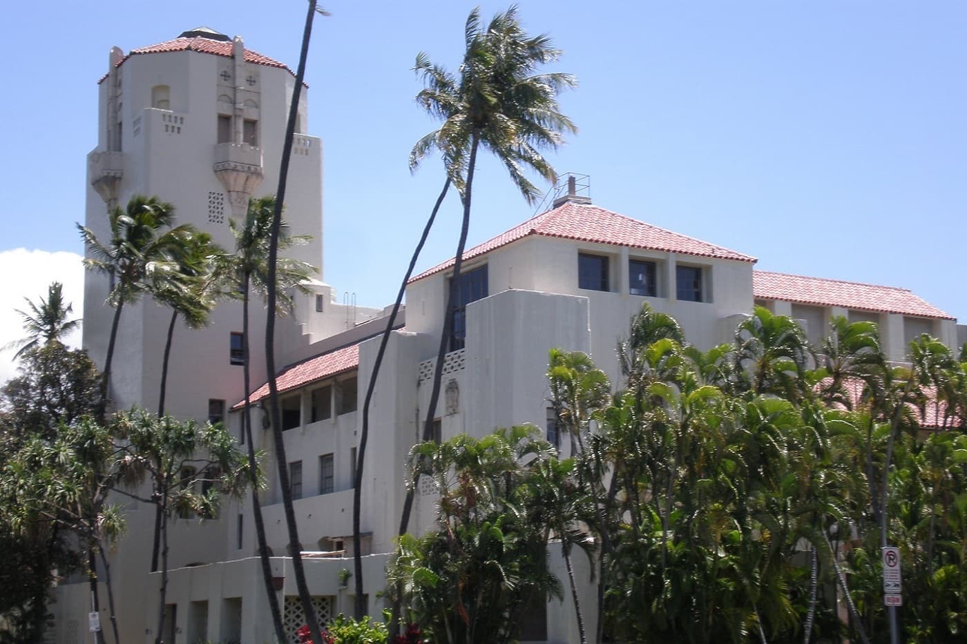 Large white building with a prominent tower and red roofs, nestled among tall palm trees.