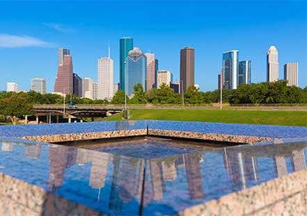Downtown Houston skyline reflected in a polished granite water feature under a clear blue sky.