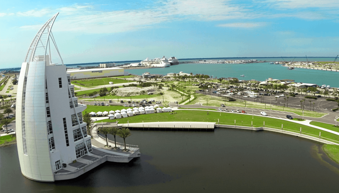 Modern sail-shaped building overlooks a busy harbor with boats and a large cruise ship.